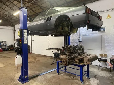 Une voiture sur un pont élévateur deux colonnes dans un atelier loué, moteur déposé sur une table roulante, établi et tableau blanc au fond.