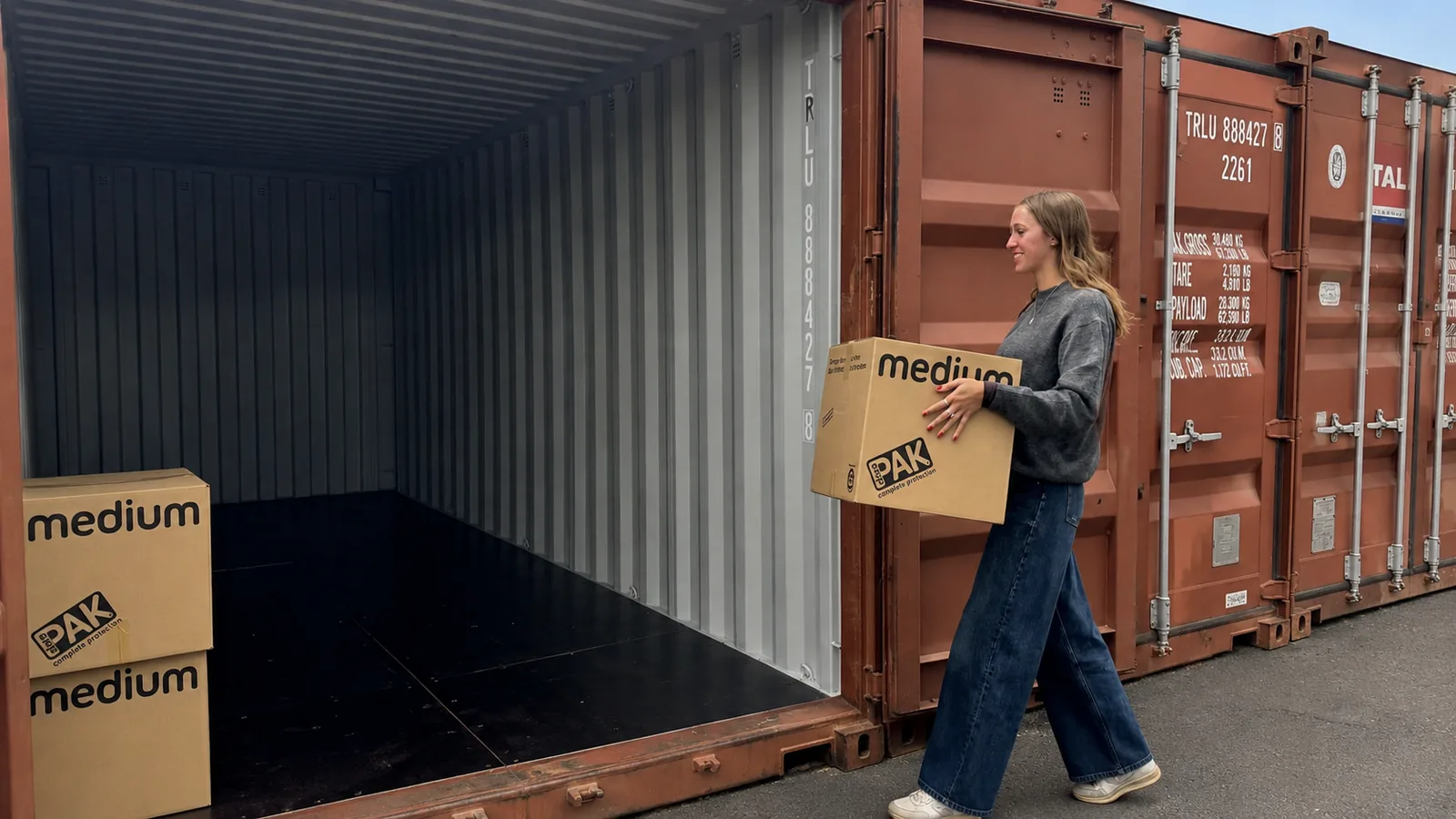 Une personne porte un carton vers un box de stockage ouvert à Givet ; l'intérieur vide se voit sur toute sa profondeur, deux autres cartons attendent au sol.