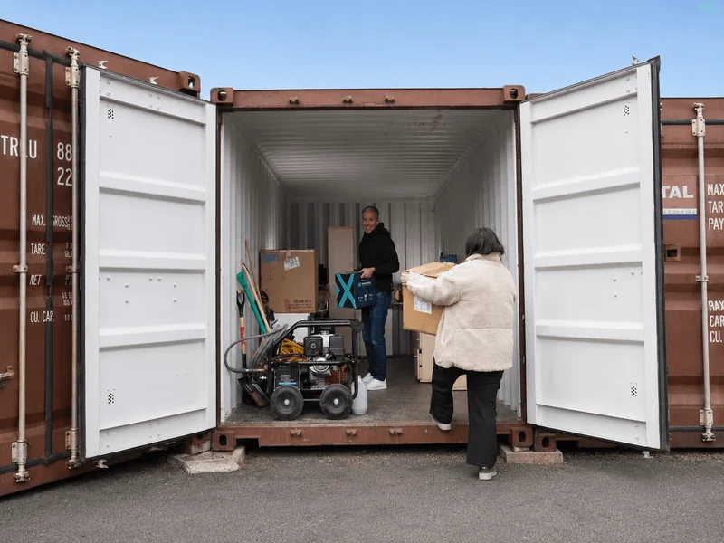 Un couple charge des cartons et une machine dans un box de stockage ouvert de plain-pied à Givet ; la profondeur du box est visible sur toute sa longueur.