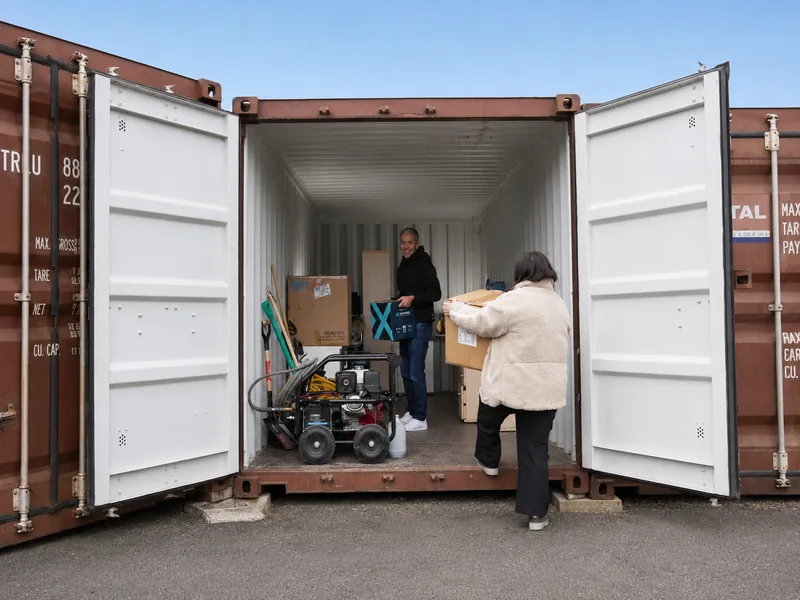 Un box de 33 m³ portes grandes ouvertes en cours de chargement : un homme debout à l'intérieur reçoit un carton que lui apporte une femme, à côté d'un nettoyeur haute pression, de pelles et de cartons déjà rangés le long de la paroi.