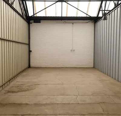 L'intérieur d'un garage vide vu depuis la porte : charpente métallique, toiture translucide qui éclaire tout le volume, mur du fond en brique peinte et prise de courant.